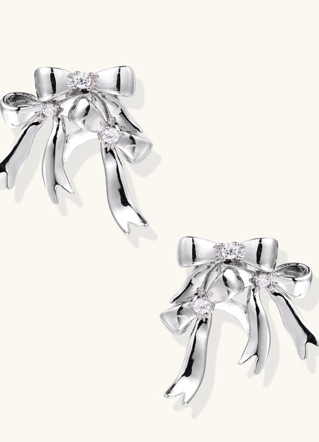 Pair of 18K gold plated brass bow-shaped earrings with flowing ribbon tails and zirconia crystal accents on white background.