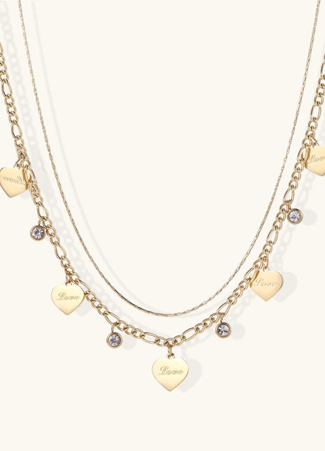 Two layered gold-plated necklaces with heart pendants engraved 'Love' and sparkling crystal charms on white background.