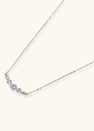 Delicate 14K gold plated necklace with curved arc of sparkling zirconia stones on cream background.