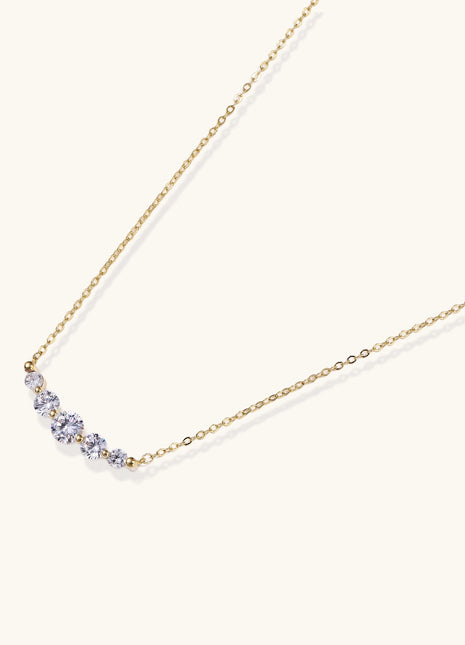 Delicate 14K gold plated necklace with curved arc of sparkling zirconia stones on cream background.