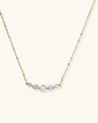 Delicate 14K gold plated necklace with curved arc of sparkling zirconia stones on cream background.