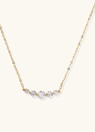 Delicate 14K gold plated necklace with curved arc of sparkling zirconia stones on cream background.